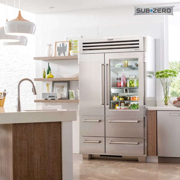 A modern kitchen with a large stainless steel Sub-Zero refrigerator, some shelves with dishes and decor, a countertop with a sink, and fresh produce on display. The refrigerator door is partly glass, showing organized food inside.